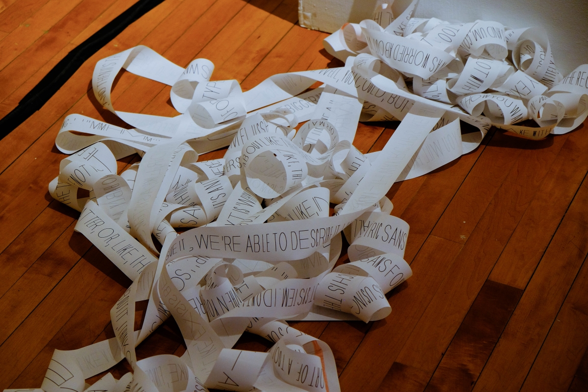 Rolls of tangled receipt paper on the ground. The text on the paper is rendered in Biometric Sans, which is a typeface that stretches the letters horizontally based on typing speed. The text is printed along the receipt paper in one infinite line.