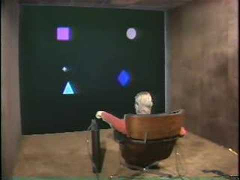An image of the Put-That-There system. A man sits in a lounge chair facing a wall. The wall has some shapes projected on it, and a cross-shaped cursor shows where the man is pointing.